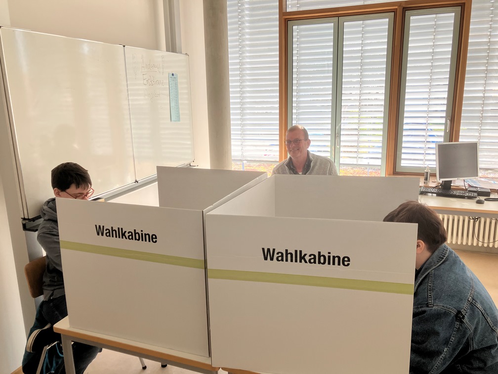 Das Foto zeigt zwei Teilnehmende in Wahlkabinen und ihren Berufsschullehrer